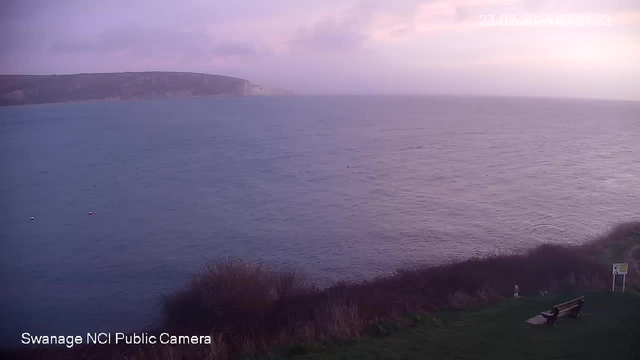 A serene view of the sea under overcast skies. The water appears calm with varying shades of blue and gray, reflecting the cloudy atmosphere. In the foreground, there is a grassy area with bushes, and a simple bench facing the water. A sign is partially visible near the bench, suggesting it is a public area. In the distance, cliffs are silhouetted against the horizon, fading into the clouds. The scene conveys a tranquil, muted environment.