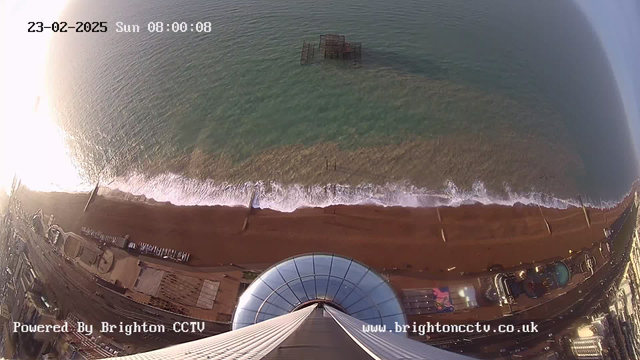 A high-angle view of a beach at sunrise, with a shoreline of golden sand meeting turquoise waves. In the foreground, a circular structure with a glass dome is visible, leading up to a tower. Beneath the water, a derelict structure can be seen, partially submerged. The scene includes elements of a boardwalk and amusement rides, suggesting a coastal area with recreational activities. The date and time are displayed in the upper left corner.