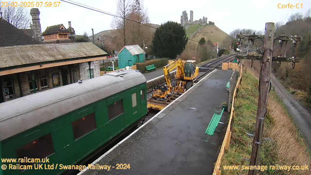 A green train carriage is parked at a railway station with a yellow construction vehicle beside it. The platform is empty, and there are several green benches along the edge. In the background, there are trees and a hill with ruins on top. The sky is overcast, and the scene appears to be early morning. There are wooden fences and a utility pole on the right side of the image.