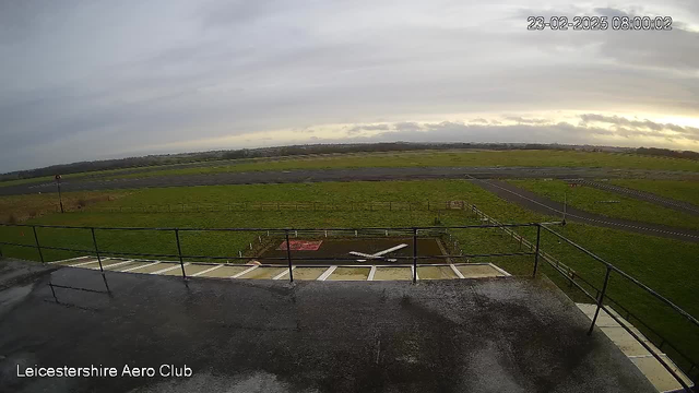 A view from a webcam at the Leicestershire Aero Club, showing a grassy field and an airstrip under a cloudy sky. In the foreground, there is a railing, and below it is an airplane marked by a white X on the ground. The airfield is bordered by fences, with open space and patches of road visible in the distance. The image appears slightly wet, suggesting recent rain.