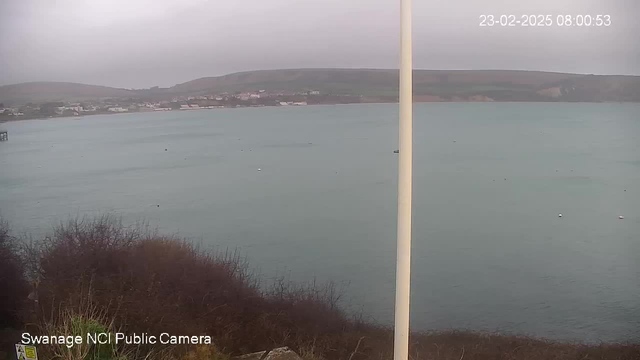 A cloudy view of a coastal area showing a calm sea with various small boats. In the foreground, there are some bushes, and a flagpole is partially visible on the right side of the image. In the background, there are buildings along the shoreline, and hills rise in the distance. The time and date display in the upper right corner reads 23-02-2025 08:00:53.