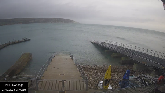 A cloudy shoreline view over a calm sea. A concrete ramp leads down to the water, flanked by a metal railing. To the left, there is a curved wooden pier extending into the water, and a rocky shoreline visible on the far left. In the foreground, several colorful kayaks—yellow and blue—are stored near the ramp. The scene is tranquil, with a gray sky and gentle waves.