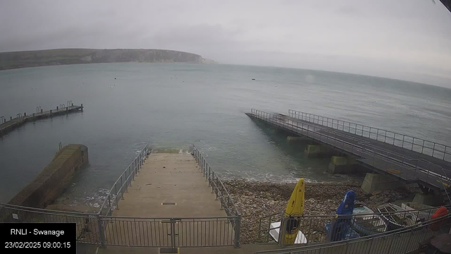 A cloudy morning view of a body of water with a rocky shoreline. There are two piers extending into the water: one is a long, narrow floating structure, while the other is a solid concrete pier. A ramp leads down from the top of a concrete platform onto the beach, which is made up of pebbles and stones. Several boats and kayaks are positioned near the water's edge, with colorful canoes in yellow, blue, and red. The horizon shows a faint outline of hills in the background.