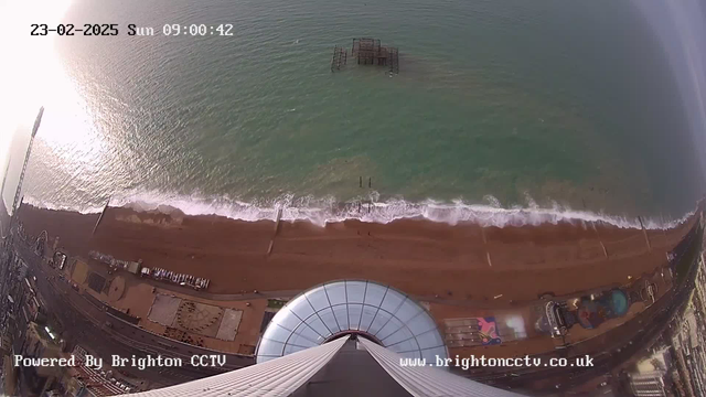 A wide aerial view of a beach on a sunny day, capturing the coastline meeting the sea. The sandy shore is visible below, with gentle waves lapping at the beach. An old, partially submerged pier is seen in the water, and various structures and attractions, like amusement rides, are lined along the beach. The image is taken from a high vantage point, showcasing the expansive blue water and shoreline. The date and time are displayed in the corner, indicating it is Sunday, February 23, 2025, at 9:00 AM.