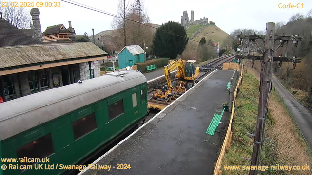 A green train carriage is parked at a station. Next to it, there is construction equipment, specifically an excavator, positioned on a flatbed. The background features a grassy hill with castle ruins at the top. The station building has a stone exterior and a roof with wooden shingles. There are several green benches on the platform, and a wooden fence runs along its edge. Power lines and a pole are visible on the right side of the image, and the scene appears to be overcast.