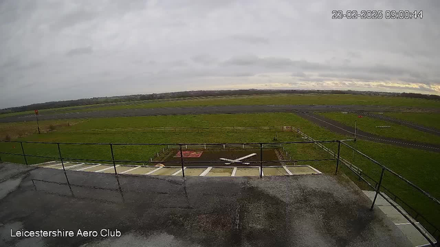 A view from a webcam showing an airfield. In the foreground, there is a wet concrete area with a railing and steps leading down. A small white airplane is positioned on the ground below, on a grassy patch marked in red. The background features a wide expanse of green grass, a gray sky with scattered clouds, and an airstrip that extends into the distance. The time and date in the corner indicate it is 09:00 on February 23, 2025.