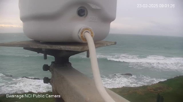 A white device mounted on a metal platform is shown in the foreground. A thick white cable extends from the device. Behind it, waves crash against rocky shoreline in a gray ocean under cloudy skies. The scene appears to be slightly overcast, suggesting a chilly atmosphere. The timestamp in the top right corner reads "23-02-2025 09:01:26," and the camera is identified as "Swanage NCI Public Camera."