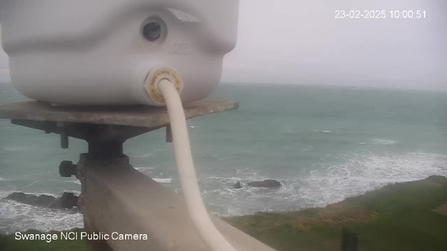 A close-up view of a white cylindrical object with a hose attached, positioned on a metallic structure. In the background, choppy turquoise water is visible, indicating the sea, with some rocky formations partially submerged. The sky is overcast, creating a dull atmosphere. The timestamp at the top indicates the date and time as February 23, 2025, at 10:00:51.