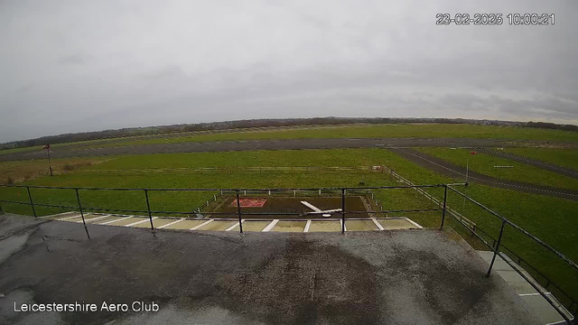 A view from above an airfield at the Leicestershire Aero Club. The surface is mostly gray with some wet areas, indicating recent rain. In the foreground, there is a railing and a concrete path leading downwards. The airfield is expansive, covered with green grass and a black asphalt runway. A windsock is visible on the right side, indicating wind direction. The sky is overcast with gray clouds, creating a somber atmosphere. A few markers and benches are scattered around the field. The date and time are displayed in the top right corner.