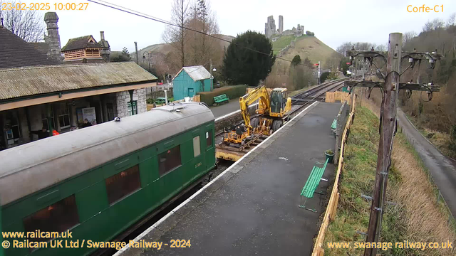 A train station scene features a green railway carriage on the left, with a yellow piece of machinery on a flatbed next to it. In the background, there are several benches painted green, and an old stone building with a peaked roof. Further back, a hill rises with the ruins of a castle on top, surrounded by trees. The sky is overcast, and there are telephone poles with wires along the right side of the image.