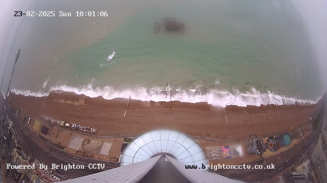 Aerial view of a beach with waves gently crashing onto the shore. The sandy beach is visible with some structures, possibly a pier, in the water. The scene is cloudy or foggy, creating a grayish atmosphere. There is a round, glass structure in the foreground at the top of the image. The date and time are displayed at the top of the image.