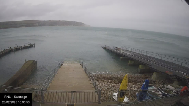 A coastal scene shows a harbor with gray waters and overcast skies. In the foreground, a wide staircase descends into the water, bordered by a railing. To the left, a curved stone jetty extends into the sea. Several small boats are visible on the shore, with a yellow kayak and a blue kayak leaning against the railing. In the distance, cliffs rise along the coastline under a cloudy sky.