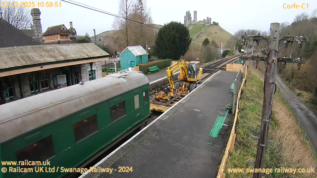 A green train carriage is partially visible on the left side of the image, with a weathered roof. In front of the train, there is a yellow construction vehicle working on the tracks. The station platform is made of dark paving stones and has several green benches along the side. Behind the platform, a stone building with a slanted roof can be seen, partially obscured by trees. In the background, a hill rises with ruins of a castle perched at the top. The sky is overcast, and there are power lines running across the image.