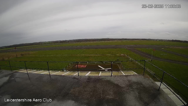 A view from a webcam mounted on a building, overlooking an airfield. The image features a green grassy area with various types of runway surfaces visible in the distance. To the left, there's a red and white windsock. The sky is overcast with gray clouds. In the foreground, there's a railing along the edge of the building. A small white object, possibly an aircraft or a glider, is located on the ground in the lower part of the image. The time and date are displayed in the top right corner.