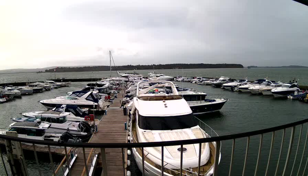 A marina scene with numerous boats docked in the water. The foreground features a large white boat with a tan deck, while smaller boats of various colors and sizes are visible in the background. There are wooden walkways connecting the boats, and the sky is overcast, suggesting a cloudy day. In the distance, a landmass is seen, indicating the shoreline. The water appears choppy, reflecting the gray sky.