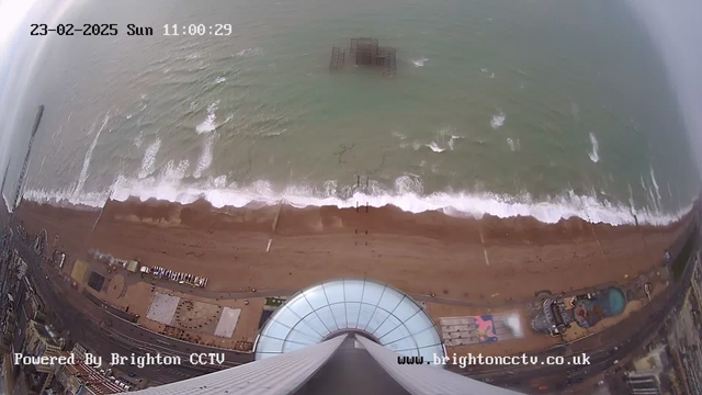 A panoramic view from a high perspective shows a sandy beach with light waves crashing against it. To the left, a pier extends into the ocean, and several small boats are visible along the shore. Beneath the viewpoint, there appears to be a circular glass structure. The scene is set on a cloudy day with limited visibility, and the date and time are displayed in the top left corner.