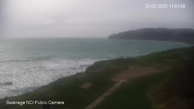 A cloudy day at the coastline of Swanage, where the turquoise waves are gently crashing against the rocky shore. The grass is lush and green, with a stone pathway leading towards the water. In the distance, a hilly landscape rises against the horizon, partially obscured by fog. The overall atmosphere is calm and serene.