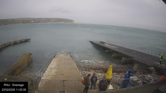 A view of a coastal area with a gray sky and calm water. In the foreground, there are three people standing at the edge of a walkway that leads down to the water. To the right, there are two boats on the shore, one yellow and one blue. A wooden jetty extends into the water, with a small rocky area visible beside it. In the background, there are cliffs and hills visible beyond the water. The scene conveys a quiet, overcast day at the seaside.