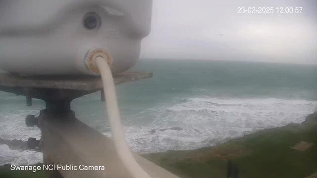 A close-up view of a white webcam mounted on a structure, with a cable attached. In the background, there is a view of a turbulent, gray ocean with white-capped waves crashing against the shoreline. The sky is overcast, creating a gloomy atmosphere. The date and time display in the corner indicates February 23, 2025, at 12:00:57. The bottom left of the image shows the text "Swanage NCI Public Camera."