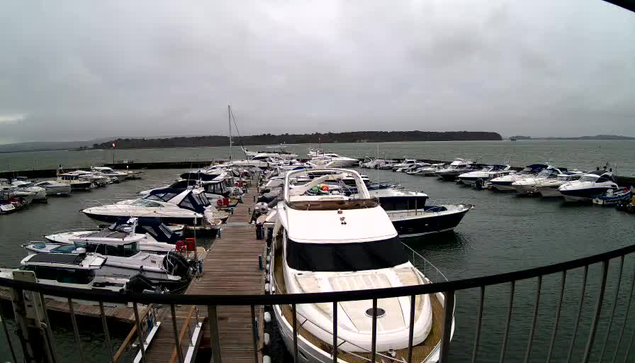 A marina filled with various boats, mostly white and blue, docked along wooden piers. The scene is set under a cloudy sky, with calm water reflecting the overcast conditions. In the background, a distant landmass is visible, and some boats are anchored near the shore.