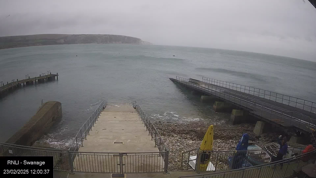 An overcast scene by the seaside with gray skies. In the foreground, there is a concrete ramp leading down to the water, flanked by a railing, and a rocky area at the base. To the right, a wooden dock extends into the water, with some boats tied up. A few people are visible near the shoreline, with a kayak and boat gear nearby. The water appears calm with gentle waves. In the background, a chalky cliff can be seen along the coast. The image is timestamped with the date and time.