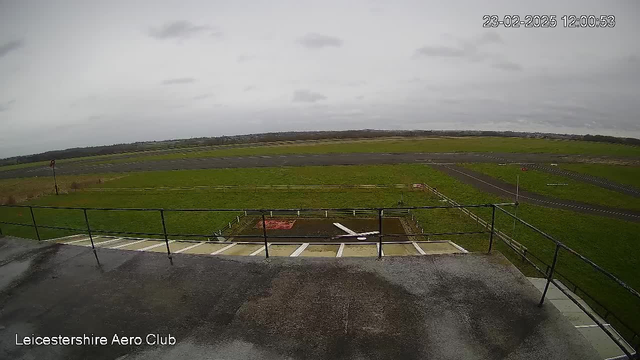 A view from a webcam shows a grassy aerodrome under a cloudy sky. In the foreground, a concrete platform with railings is visible, leading down to an area with a model airplane placed on the ground. An asphalt runway stretches into the distance, surrounded by green fields and a few scattered trees. There are some signs and a wind indicator visible on poles near the runway. The atmosphere appears overcast and calm.