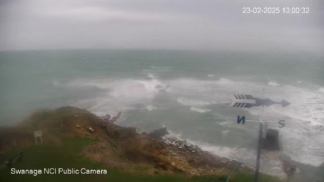 A turbulent sea under a gray sky, with large waves crashing against rocky cliffs. The scene is windy, causing spray to rise above the water. In the foreground, there is vegetation and a bench on the grassy area. A weather vane indicates direction, with letters "N" for north and "S" for south visible. The image is timestamped with "23-02-2025 13:00:32" in the corner.