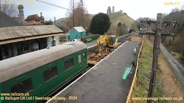 A view of a railway station at Corfe, featuring a vintage green train on the left side. There is a yellow excavator machine positioned next to the tracks. In the background, a large grassy hill rises, topped with a historic castle. The station building, with stone walls and a tiled roof, is partially visible. Several benches line the platform, and trees are scattered around the area. The scene is set under a cloudy sky, with electrical poles and wires adding to the railway setting.