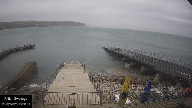 A cloudy day by the sea, showing a view of a concrete ramp leading down to water. The ramp is flanked by metal railings and leads to a rocky shoreline. To the left, a small pier extends out into the water, and another larger pier is visible on the right, with railings and stairs. Some boats are moored nearby, and in the foreground, there are colorful kayaks secured near the edge. The sea is calm with gentle waves lapping against the shore.