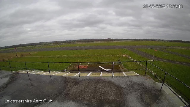 A view from a balcony overlooking an airfield on a cloudy day. The foreground shows a flat surface with railings, and a partial view of a white structure on the ground. The background features a large green grass area with a runway marked by gray pavement. There are some small structures and signs in the distance, and the sky is overcast. The image is timestamped and identifies the location as Leicestershire Aero Club.