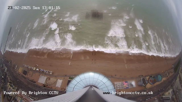 A panoramic view of a beach and ocean taken from a high vantage point. The scene shows rolling waves crashing against the sandy beach below, with a pier visible in the distance. The beach has scattered people enjoying the day. Various structures can be seen along the shoreline, including a circular design in the sand and buildings with colorful attractions. The sky is overcast, and the time and date are displayed in the top left corner.