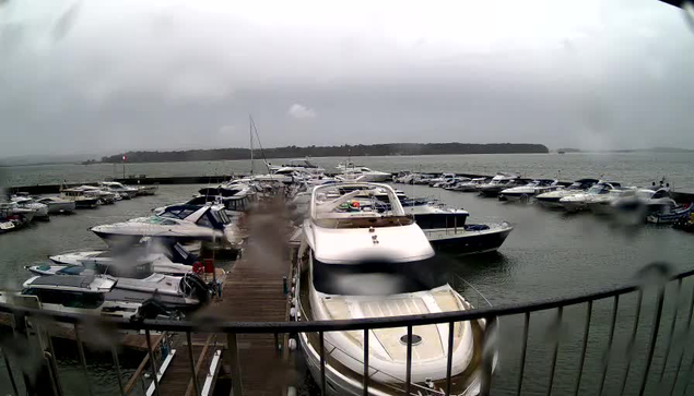 A view of a marina on a cloudy day with rain droplets on the camera lens. Multiple boats and yachts are moored along the dock, with some larger vessels in the foreground. The water appears choppy, and there is a distant shoreline with trees visible in the background. The scene gives a sense of a tranquil yet overcast environment.