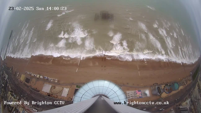 A webcam image showing a bird's-eye view of a beach with waves crashing over sandy shorelines. The water is a muted blue-grey color, with whitecaps visible where the waves break. In the background, a pier extends into the water, partially submerged and obscured by waves. Various shapes and colors of beach structures and equipment can be seen along the shore, including a circular design on the sand. The image features a clear sky and digital timestamps indicating the date and time.