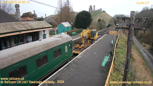 A view of Corfe Castle railway station showing a green train car parked at the platform. On the right, a yellow construction vehicle is positioned next to two tracks, with tools attached for track maintenance. The station building is visible in the background, with several benches along the platform, and green sheds and trees nearby. In the distance, the remains of Corfe Castle can be seen atop a hill. The weather is overcast and it is daytime.