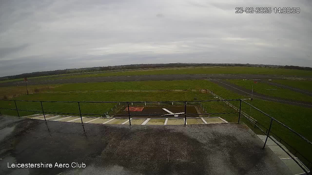 A view from a webcam positioned at the Leicestershire Aero Club. In the foreground, there is a flat rooftop area with a railing. Below, a grassy field dominates the scene, with a section of the field resembling a small runway. The sky is overcast, and the lighting is soft, suggesting a cloudy day. Parts of the runway are visible in the distance, bordered by grass and a few markings, possibly indicating a landing strip. There are no visible aircraft in the frame.