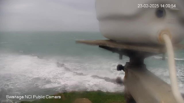 A coastal view showing turbulent waves crashing against rocky shoreline. The camera is slightly out of focus, with a circular object and cable in the foreground. The sky is overcast, contributing to a grey, gloomy atmosphere. The timestamp in the corner indicates it is February 23, 2025, at 2:00 PM.