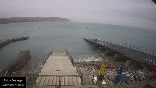 A view of a seaside area featuring a cloudy sky and choppy water. In the foreground, there are steps leading down to a rocky shoreline. To the left, a curved wooden pier juts out into the water, while a straight, wider pier is visible on the right. There are some boats secured along the pier, with a yellow kayak and other equipment nearby on the ground. The overall atmosphere is overcast, indicative of a windy day.