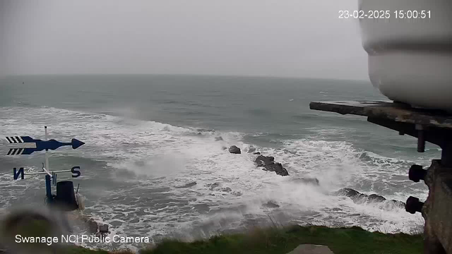 A cloudy day at a coastal location, with waves crashing against rocky outcrops in the foreground. In the lower left corner, there is a directional sign with arrows indicating north and south. The setting appears stormy, with a grey sky and rough sea, and the image has a timestamp of February 23, 2025. There are blurred elements in the lower corner, possibly indicating a camera housing.