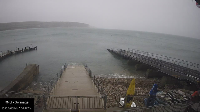 A view of a coastal area with a foggy atmosphere. In the foreground, there is a concrete pathway leading to the water, bordered by railings. To the left, there is a small pier with several posts. On the right, another wooden pier extends out over the water. The sea appears calm, with faint ripples, and rocky terrain is visible along the shore. The background features a misty landscape with hills partially obscured by fog. The date and time are displayed in the bottom left corner.