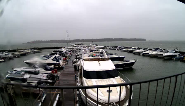 A waterfront scene depicting a marina filled with various boats parked at docks. The sky is overcast and appears gray, suggesting possible rain. The surface of the water is calm but slightly rippled. A wooden walkway can be seen in the foreground, leading to the boats. The area appears quiet, with several boats of different sizes, some with white hulls and others in darker colors, lined up along the docks.