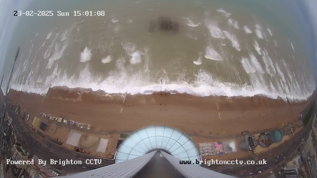 An aerial view of a beach on a cloudy day, showing waves crashing onto the shore. In the foreground, there is a circular observation platform, while the sandy beach stretches out below. Various beachside structures and amusement rides are visible along the coast, with some people walking along the shore. The date and time are displayed in the upper left corner.