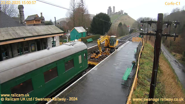 A green train carriage is parked on a railway platform. An excavator is positioned next to the train, performing maintenance work on the tracks. The setting is overcast with a slight drizzle, and the platform is wet. In the background, the remains of a castle are visible on a hill, surrounded by trees. There are some benches and a small green building on the platform, with a wooden fence and power lines nearby.