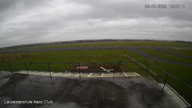 A view from a webcam at the Leicestershire Aero Club. The foreground shows a flat, wet surface with a railing, leading to an open field. The field is primarily green grass with a few patches of gray pavement in the background. There are two distinct areas of gray pavement on the far side, possibly runways, and a red warning flag on a pole. The sky is overcast with gray clouds, indicating a cloudy day.