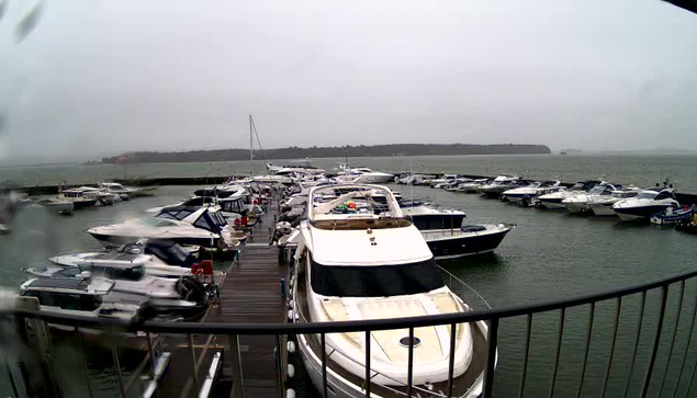 A marina filled with various boats docked at a pier. The sky is overcast and gray, indicating cloudy weather. The water appears choppy, reflecting the gloomy atmosphere. There are several boats of different sizes and designs, with some moored close together. In the foreground, a larger white yacht is visible with a black canopy. The scene is framed by a railing in the lower part of the image.