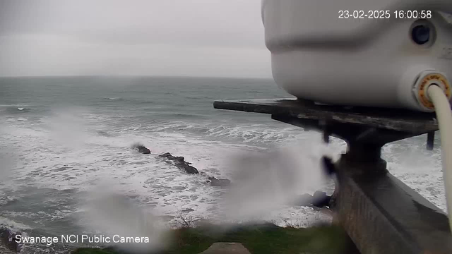 A cloudy scene over the ocean, with gray skies and rough waves crashing against dark rocks in the water. The image is partially obscured by water droplets on the lens, making it difficult to see clearly. In the foreground, part of a white camera or device is visible, resting on a gray ledge. The timestamp at the top indicates the date and time.