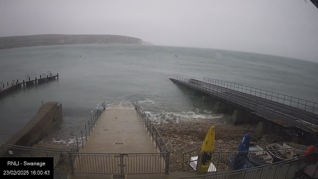 A coastal scene depicting a gray, overcast day. In the foreground, there is a set of concrete steps leading down to a rocky beach, surrounded by metal railings. To the left, a boat ramp extends into the water. There are several small boats and bright yellow kayaks on the shoreline. In the background, the sea appears choppy with waves lapping at the shore. A distant cliff is visible on the horizon, partially obscured by mist. The timestamp at the bottom indicates the date and time.