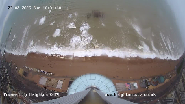 Aerial view of a beach with waves crashing onto the shore. The sand is visible, with a few structures and people scattered along the beach. A large glass structure is seen in the foreground, leading down from what appears to be a tall building. The ocean is a murky greenish-brown color, and there are white foamy areas where the waves break. In the distance, a weathered pier structure is partially submerged in the water. The scene is captured on February 23, 2025, at 16:01.