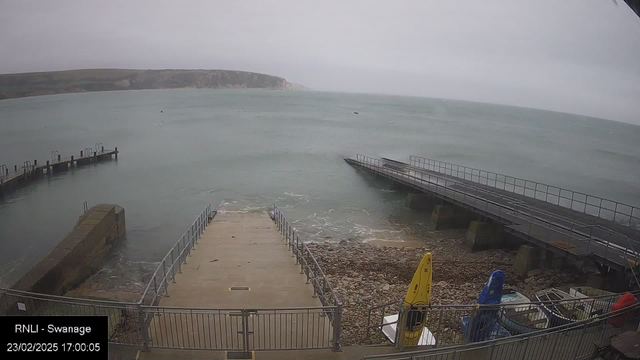 A view of a cloudy seaside location with gentle waves. In the foreground, there is a concrete ramp leading down to the water, bordered by a metal railing. On the left, a small wooden pier extends into the water, with posts visible along its edge. On the right, a larger wooden structure also juts out into the sea. The shoreline features rocks surrounding the water, and several small boats are visible near the edge, with one yellow kayak and blue equipment leaning against the railing. The background showcases a rocky cliff under overcast skies.