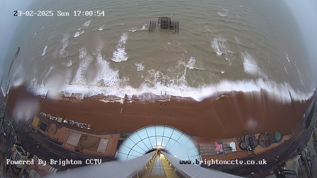 Aerial view from a high webcam overlooking a beach. Brown sand is visible at the bottom of the image, while the ocean occupies the upper part. Waves crash onto the shore, with white foam visible. A pier structure with some missing sections is located in the water, slightly off-center. Below, various shapes and colors hint at beachside facilities. The scene is cloudy and appears to be taken in the late afternoon, indicated by the timestamp in the corner.