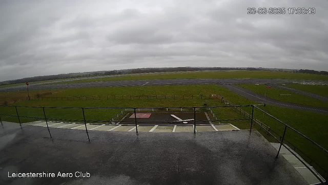 A view from a webcam showing a cloudy sky above a grassy area and an airfield. In the foreground, there is a railing and a smooth surface with some water. In the distance, the runway is visible with a few parked vehicles along the edge. The landscape is flat and green, surrounded by a fence. The image captures a gloomy, overcast day.